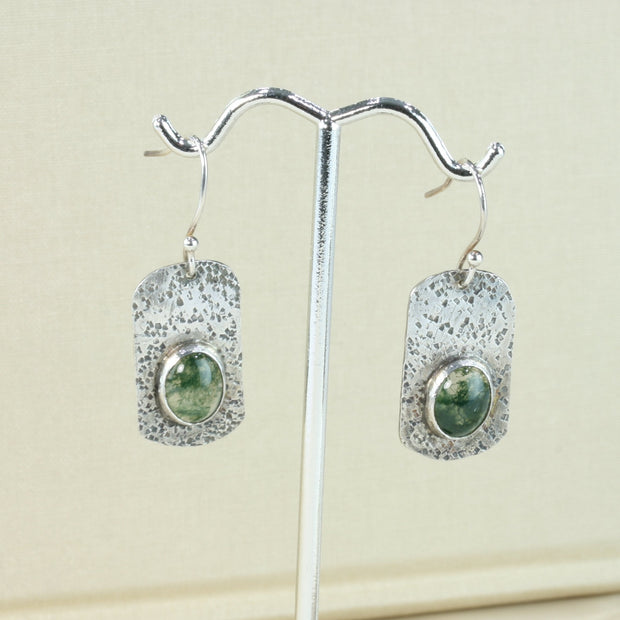 Silver hook earrings featuring a rectangle shape with a round hammered texture. The silver has been oxidised to give it a darker finish. The Moss Agate gemstones are oval and the back behind them has been cut out to let the light shine through , perfectly showing the details of the gemstones.