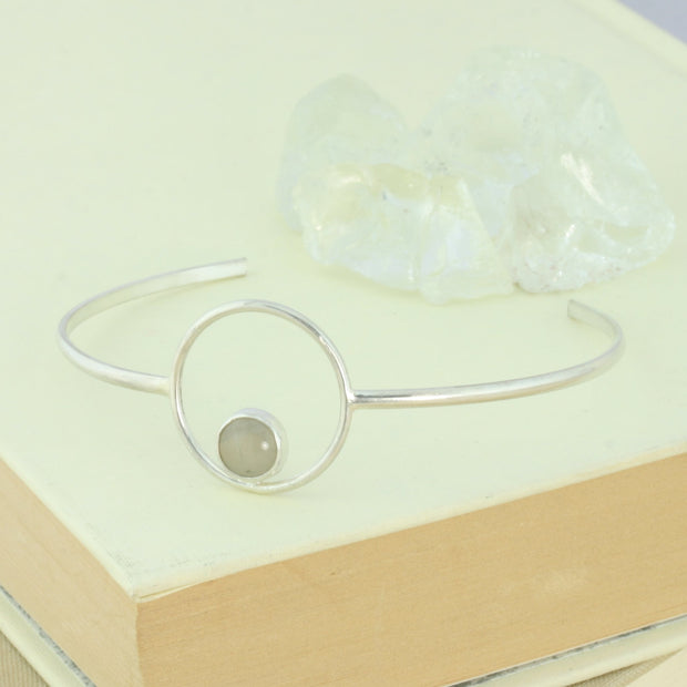 Silver bangle bracelet featuring a hoop in the centre. The hoop features a gemstone in the bottom centre, the gemstone is 8mm in diameter. This bangle features a Grey Moonstone. The silver is polished to a shiny matte finish.