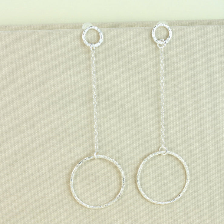 Silver earrings featuring a small hoop at the top with a stud attached to go through the ear. Chain is attached to the small hoop with a jump ring. At the bottom a larger hoop is featured. The hoops have a hammered texture and a sparky shiny finish. A matching y necklace is available.
