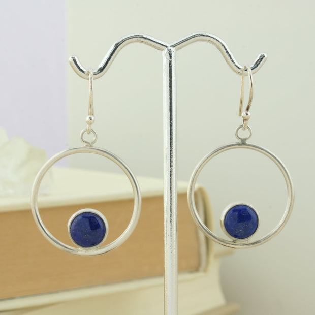 Silver hoop hook earrings featuring a Lapis Lazul gemstone. The gemstone is 8mm in diameter and is set in a cabochon setting at the bottom on the inside of the hoop. These are handmade earrings, made form eco-silver.