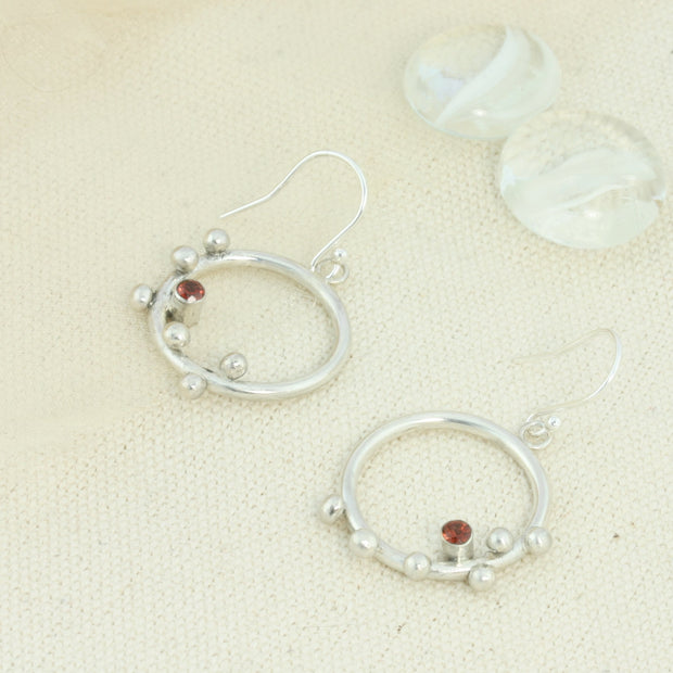 Silver hoop hook earrings featuring 6 silver balls and a Garnet gemstone. The gemstone is 3mm in diameter and is set in a tube setting at the bottom and slightly to one side on the inside of the hoop. These are handmade earrings, made form eco-silver.