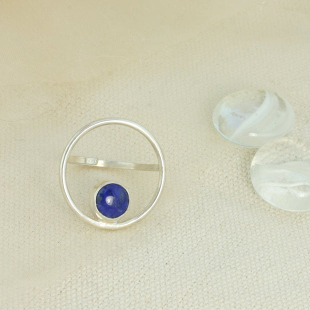 Silver ring featuring a hoop which is 2cm in diameter. The ring band is attached on one side of the ring, leaving the other side open and slightly adjustable. The hoop features a cabochon gemstone which is 8mm in diameter. This ring features a Lapis Lazul gemstone. 