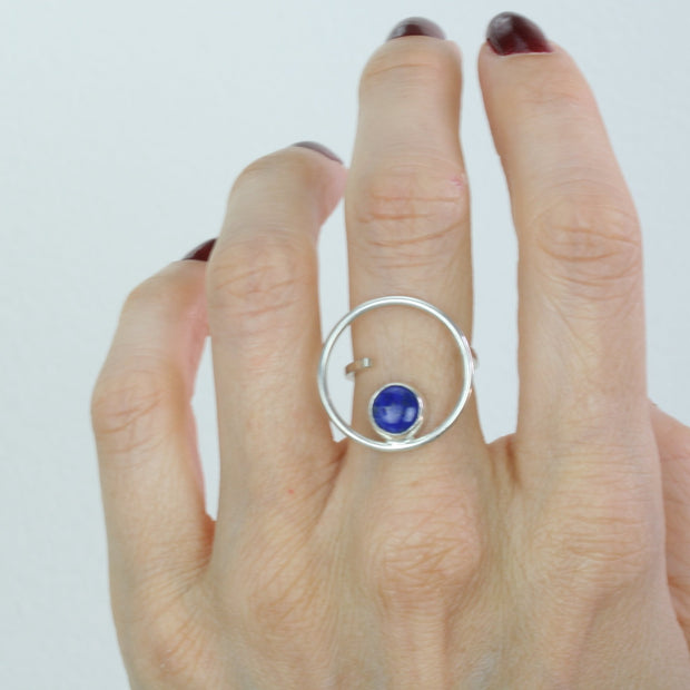 Silver ring featuring a hoop which is 2cm in diameter. The ring band is attached on one side of the ring, leaving the other side open and slightly adjustable. The hoop features a cabochon gemstone which is 8mm in diameter. This ring features a Lapis Lazul gemstone.