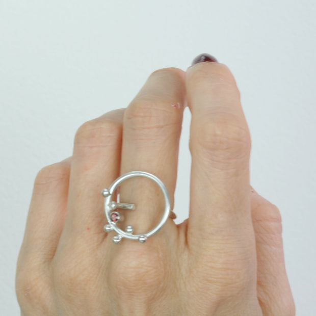 Eco silver ring featuring a hoop of 2cm in diameter. With 6 silver balls at the top, and a Garnet gemstone in the middle. The gemstone is 3mm in diameter and the ring has a slight matte finish. The band is open and attached to one side of the hoop, this makes the ring slightly flexible in size. Shown here with the matching hook earrings.