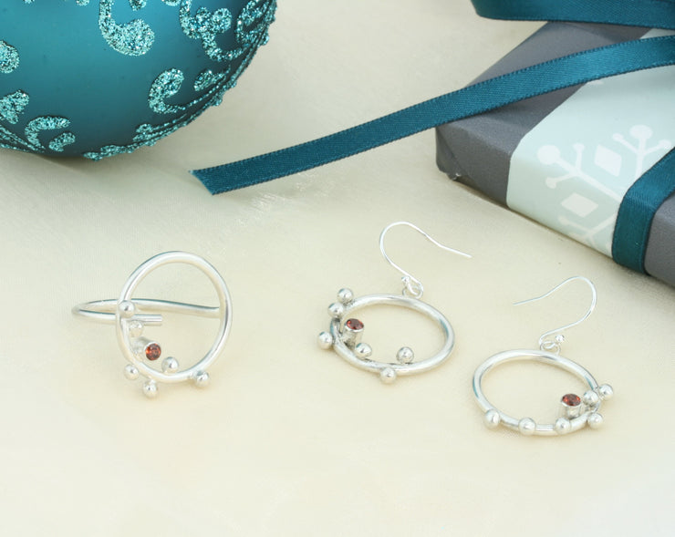 Silver hoop hook earrings featuring 6 silver balls and a Garnet gemstone. The gemstone is 3mm in diameter and is set in a tube setting at the bottom and slightly to one side on the inside of the hoop. These are handmade earrings, made form eco-silver. Shown here with the matching ring.