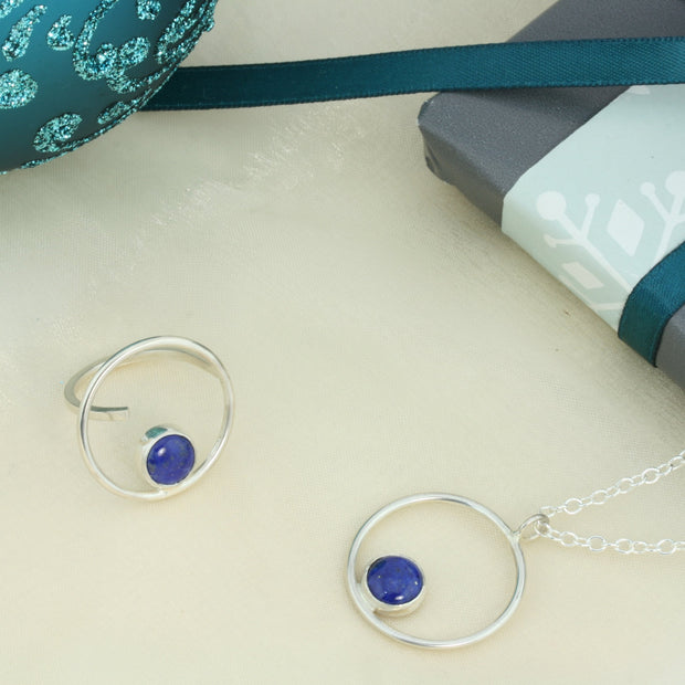 Silver ring featuring a hoop which is 2cm in diameter. The ring band is attached on one side of the ring, leaving the other side open and slightly adjustable. The hoop features a cabochon gemstone which is 8mm in diameter. This ring features a Lapis Lazul gemstone. Shown here with the matching pendant necklace.