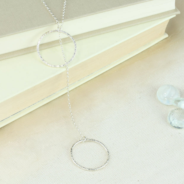 Silver y necklace featuring a hoop at either end of the chain. The hoops have been flattened and given a hammered texture and shiny finish. The chain loops through one of the hoops. By pulling the lower hoop you can adjust how high or low it sits.