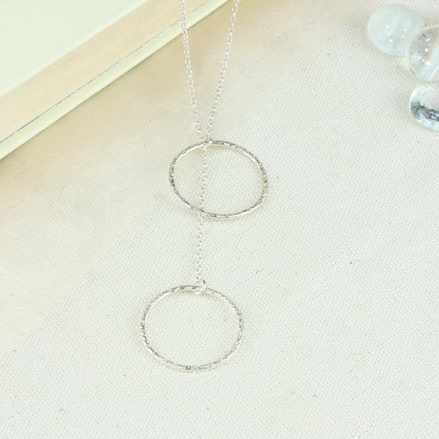 Silver y necklace featuring a hoop at either end of the chain. The hoops have been flattened and given a hammered texture and shiny finish. The chain loops through one of the hoops. By pulling the lower hoop you can adjust how high or low it sits.