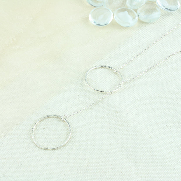 Silver y necklace featuring a hoop at either end of the chain. The hoops have been flattened and given a hammered texture and shiny finish. The chain loops through one of the hoops. By pulling the lower hoop you can adjust how high or low it sits.