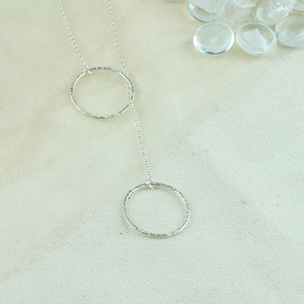 Silver y necklace featuring a hoop at either end of the chain. The hoops have been flattened and given a hammered texture and shiny finish. The chain loops through one of the hoops. By pulling the lower hoop you can adjust how high or low it sits.