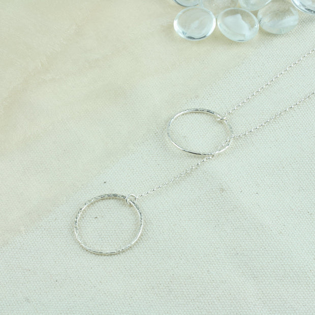 Silver y necklace featuring a hoop at either end of the chain. The hoops have been flattened and given a hammered texture and shiny finish. The chain loops through one of the hoops. By pulling the lower hoop you can adjust how high or low it sits.