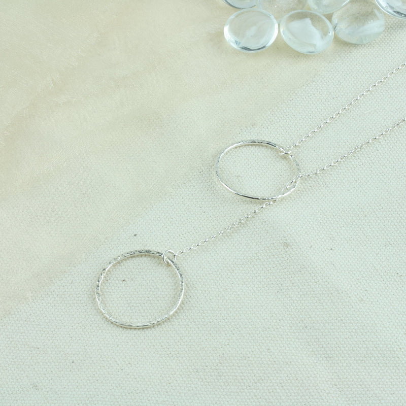 Silver y necklace featuring a hoop at either end of the chain. The hoops have been flattened and given a hammered texture and shiny finish. The chain loops through one of the hoops. By pulling the lower hoop you can adjust how high or low it sits.