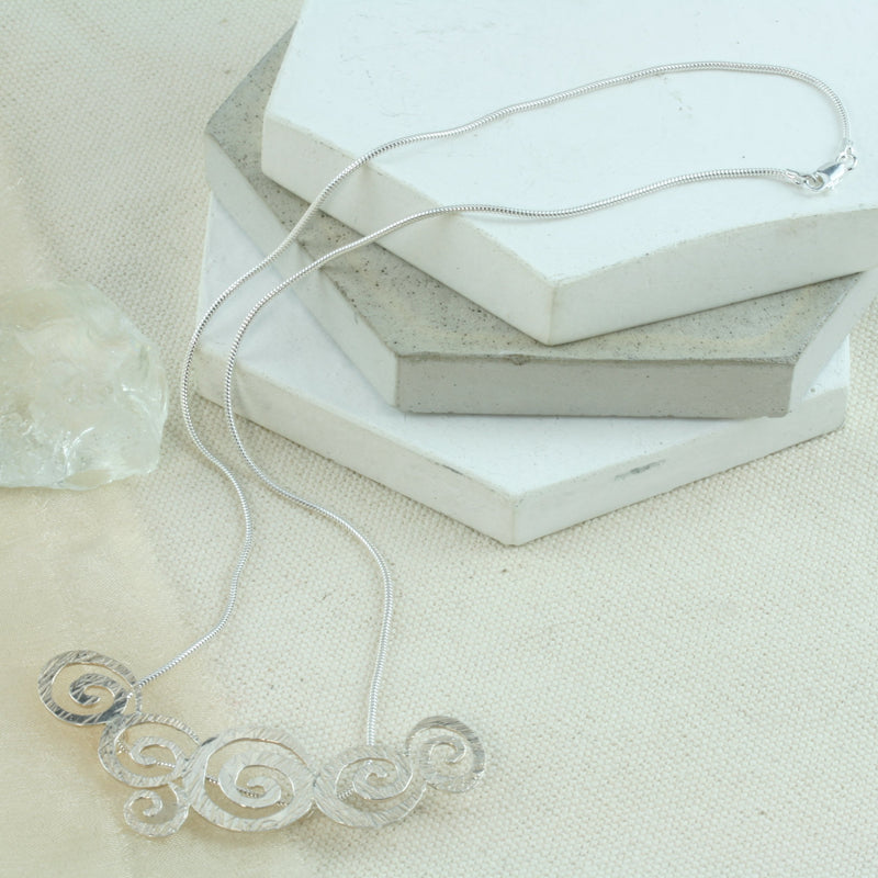 Silver necklace featuring a pendant with 6 different sized swirls. They all have a striped texture all completed with a snake chain to feature them.