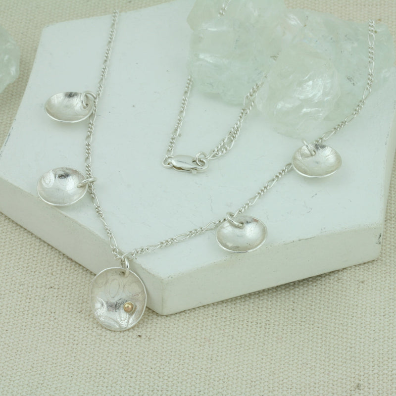 Silver necklace with 5 domed cups. There are 4 smaller cups and one larger cup which features a 9ct gold ball. The cups have a pebble texture and a mirror finish.