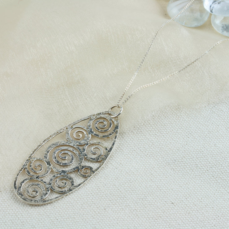 A large oval pendant necklace featuring 8 different sizes swirls all around the inside of the oval frame. It has a round hammered texture and a shiny finish which captures and reflects the light to make the necklace sparkle. 