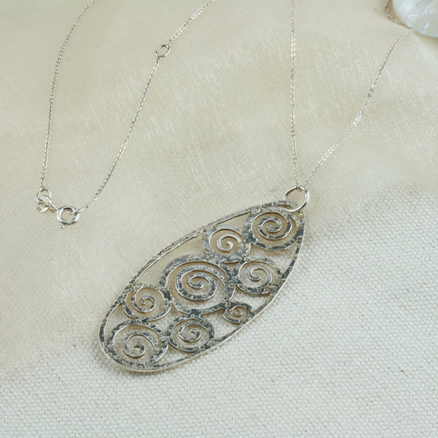 A large oval pendant necklace featuring 8 different sizes swirls all around the inside of the oval frame. It has a round hammered texture and a shiny finish which captures and reflects the light to make the necklace sparkle. 