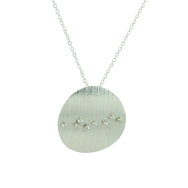 Eco silver circle pendant with a striped texture. 8 silver balls are placed in a slight diagonal line at random spots from left to right. The circle is slightly arched and has a mirror finish.