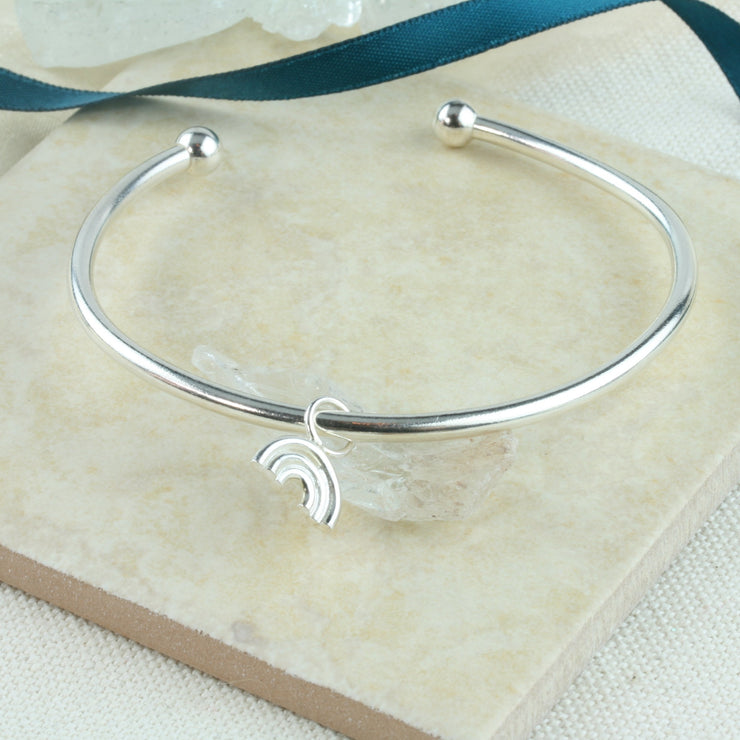 Eco silver bangle with a small rainbow charm. It is attached to the bangle with a jump ring, so it moves freely around the bangle. The bangle has a silver ball on each end and can be adjusted in width by pulling or pinching it gently. The rainbow features three arches made from round eco silver wire. The whole piece has a shiny mirror finish.