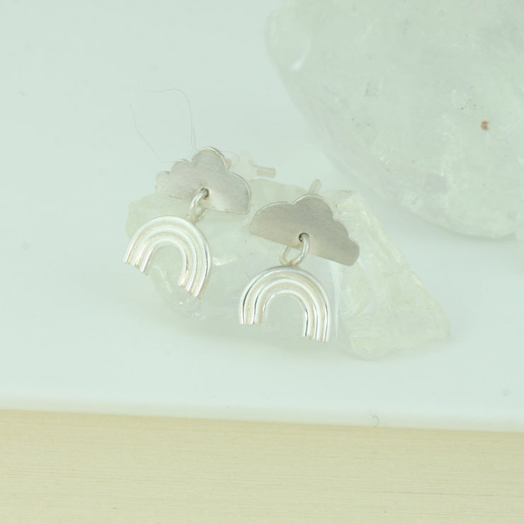 Eco silver stud earrings featuring a cloud shape with a rainbow underneath. The rainbow is attached to the cloud by a jump ring and dangles down. The rainbow has three arches made from round silver wire