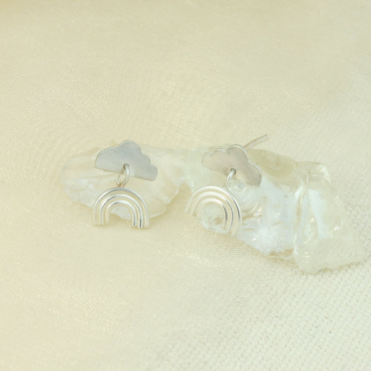 Eco silver stud earrings featuring a cloud shape with a rainbow underneath. The rainbow is attached to the cloud by a jump ring and dangles down. The rainbow has three arches made from round silver wire