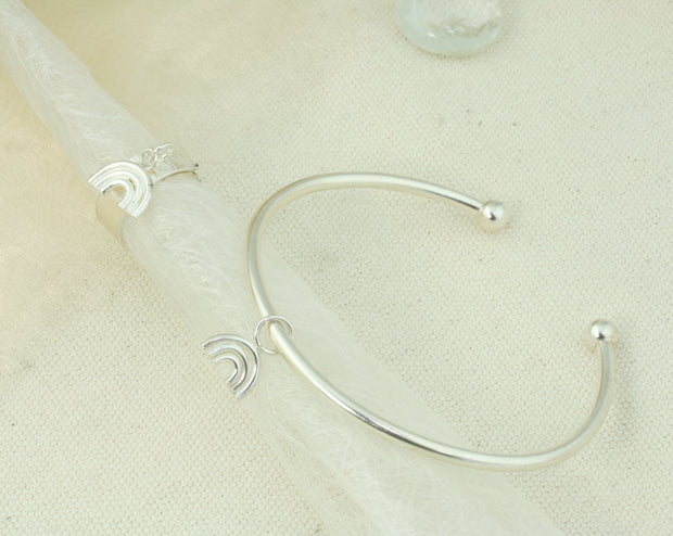 Eco silver bangle with a small rainbow charm. It is attached to the bangle with a jump ring, so it moves freely around the bangle. The bangle has a silver ball on each end and can be adjusted in width by pulling or pinching it gently. The rainbow features three arches made from round eco silver wire. The whole piece has a shiny mirror finish. Shown here with a matching adjustable ring with a rainbow charm.