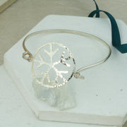 Silver bangle featuring a snowflake in the front in the middle. The snowflake shape is framed in a round border which holds both sides of the bangle in place. One side slides in and out of the jump ring to put the bangle on or take it off. The snowflake and frame have a hammered texture and shiny finish. The bangle has a mirror finish.