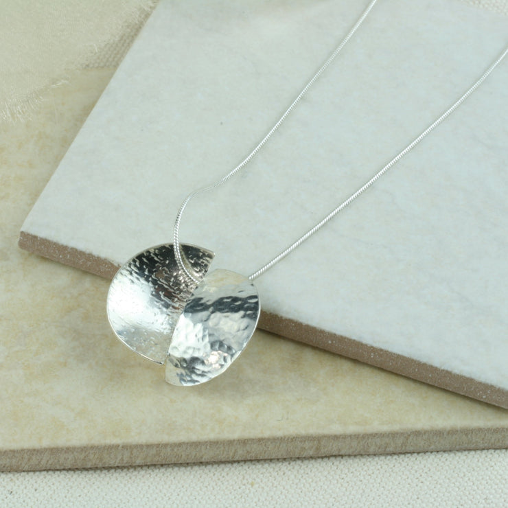 Silver pendant necklace featuring a circle that has been cut in half. Both sides have been domed into half a cup shape and soldered together with the dome on different sides. One side has a striped texture the other a round hammered texture. It has an mirror finish. The snake chain necklace  is 40 cm / 15" long.