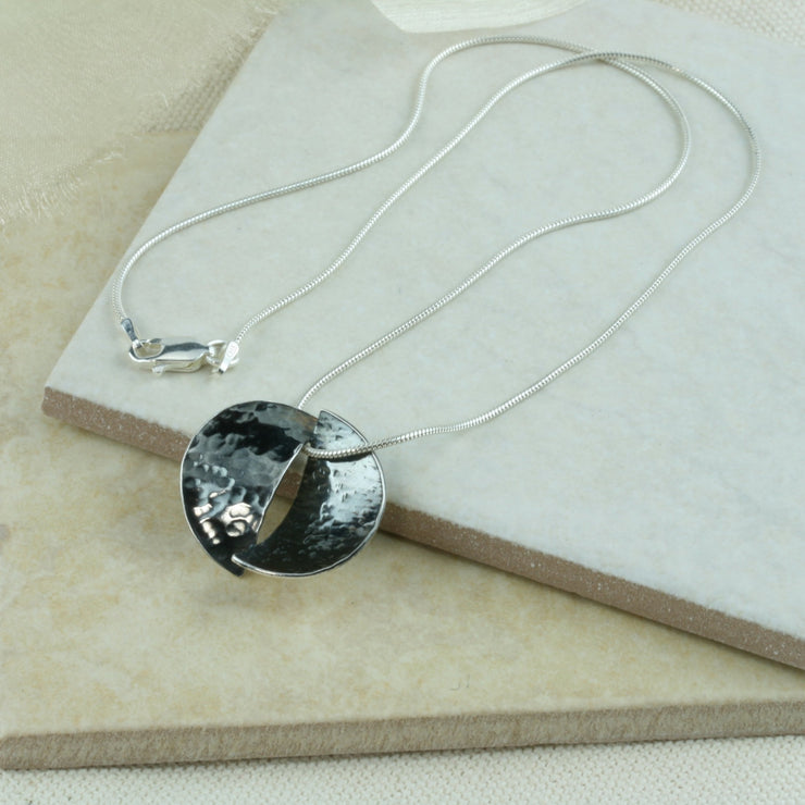 Silver pendant necklace featuring a circle that has been cut in half. Both sides have been domed into half a cup shape and soldered together with the dome on different sides. One side has a striped texture the other a round hammered texture. It has an oxidised darker finish. The snake chain necklace  is 45 cm / 18" long.