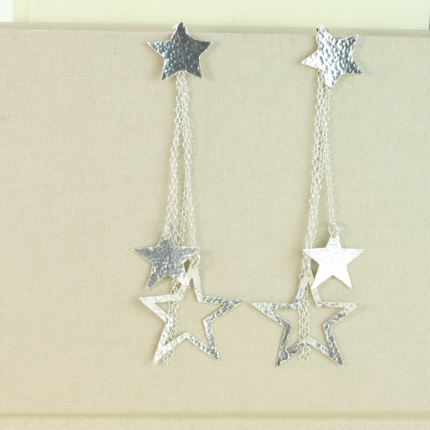 Silver star earrings featuring star studs and a separate drop part which is looped through the stud at the back. The drop features 4 chain parts with a smaller closed star and a larger open star attached to two of the chains. They have a hammered shiny finish at the front and a mirror finish at the back.