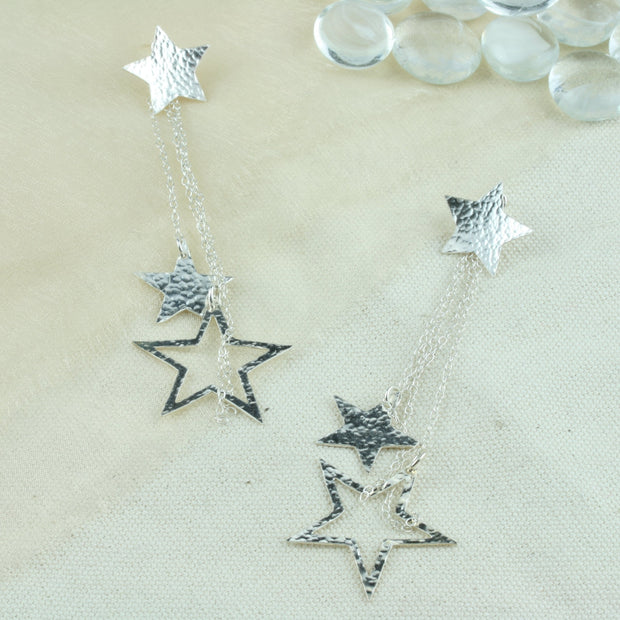 Silver star earrings featuring star studs and a separate drop part which is looped through the stud at the back. The drop features 4 chain parts with a smaller closed star and a larger open star attached to two of the chains. They have a hammered shiny finish at the front and a mirror finish at the back.