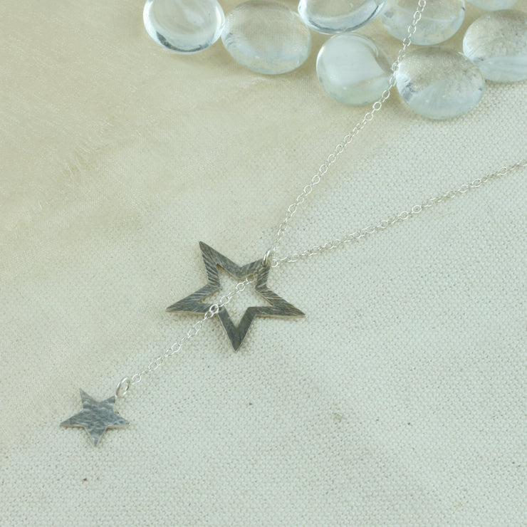 Silver necklace featuring a smaller stars on one end and a larger star on the other. The chain is looped through the larger star. Both stars have a textured finish, the larger star stripes and and the smaller star round dots.  Their backs have a mirror finish. By pulling the smaller star through the larger star you can adjust the length.