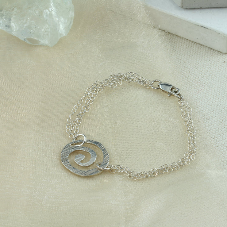 Silver chain bracelet with three chains and a silver swirl in the middle. The swirl has a striped hammered texture and a shiny finish.