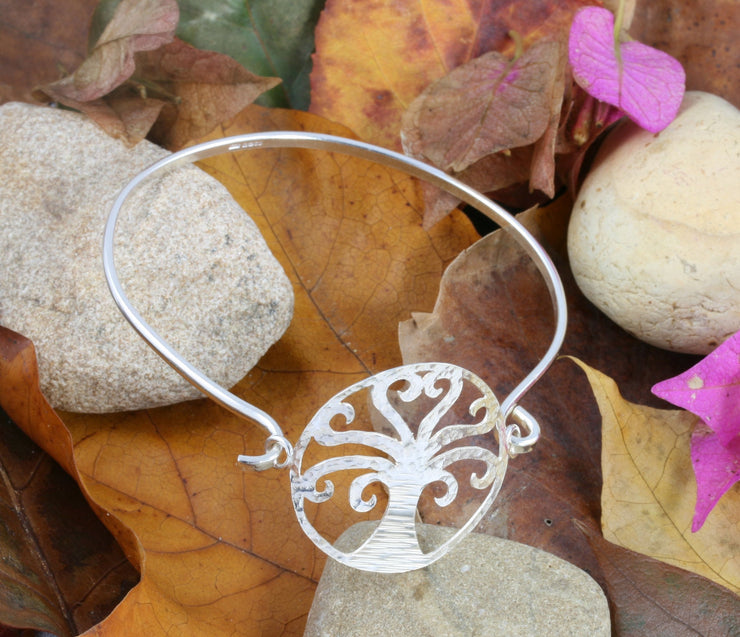 Silver bangle bracelet featuring a TRee of Life at the front. The wire of the bangle is fastened on one side of the Tree of Life. The other side can be looped through the jump ring attached to the Tree of Life to open and close the bangle bracelet. It is made from eco silver and has a shiny finish. The Tree of Life has a hammered texture.